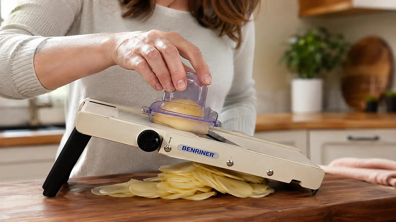 Hand guiding a potato across a mandoline slicer using the finger guard, thin even slices falling below